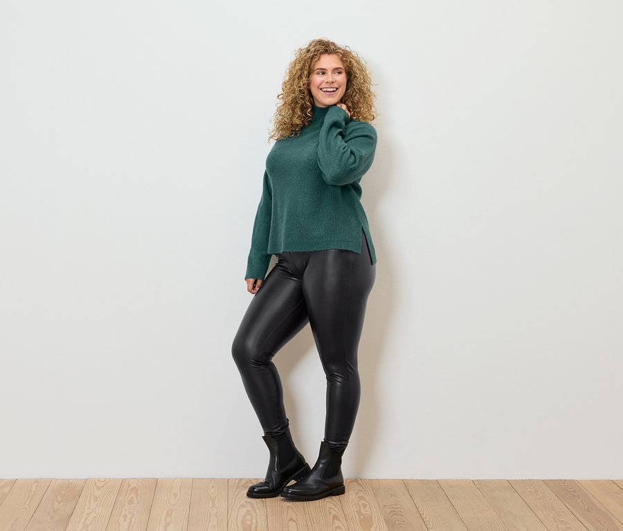 Frau mit lockigem Haar posiert in einem grünen Rollkragenpullover, schwarzen Kunstlederleggings und schwarzen Stiefeln.