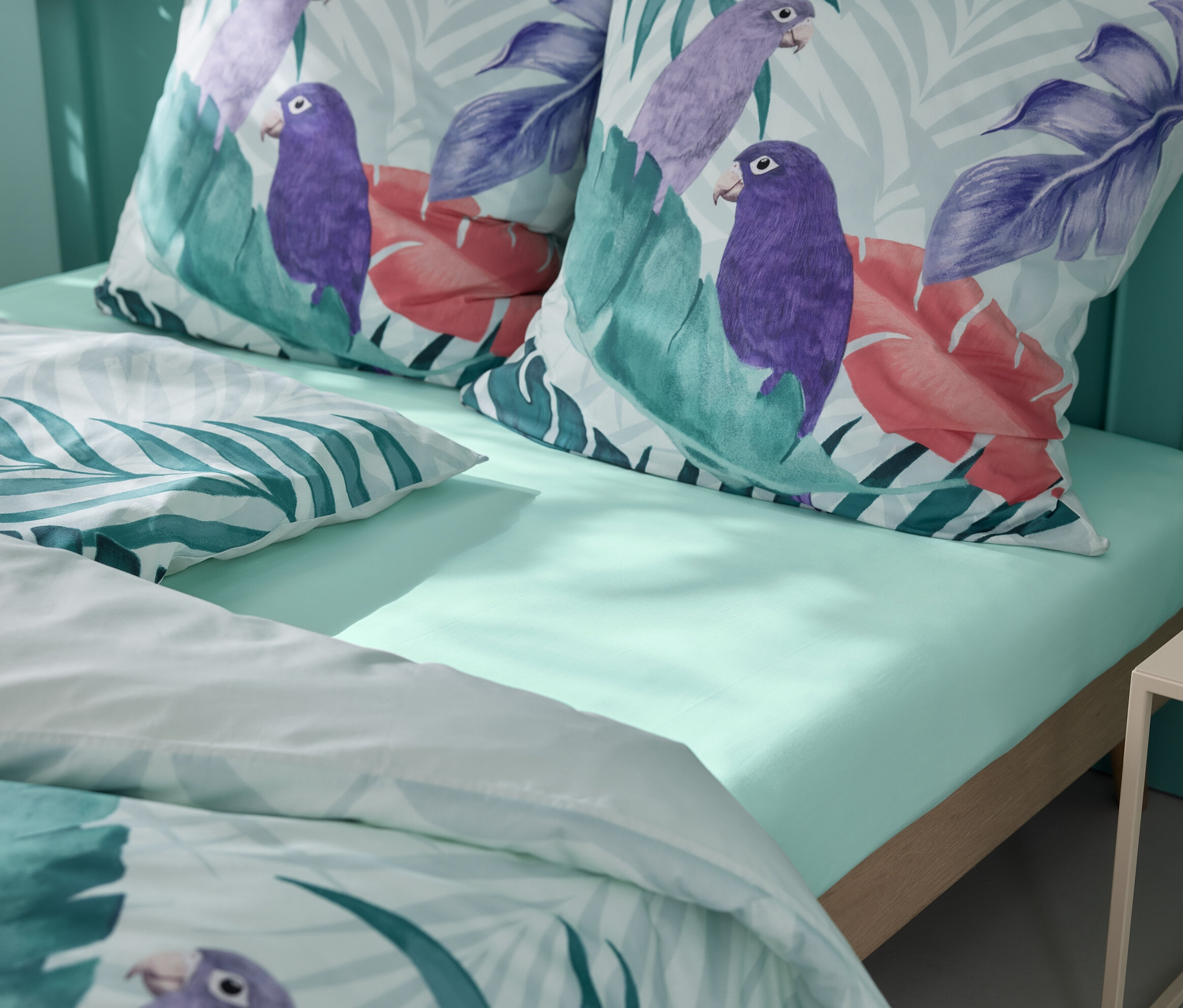 Hellgrünes Spannbettlaken mit zwei Kissen mit Papageien- und tropischen Blättermustern.