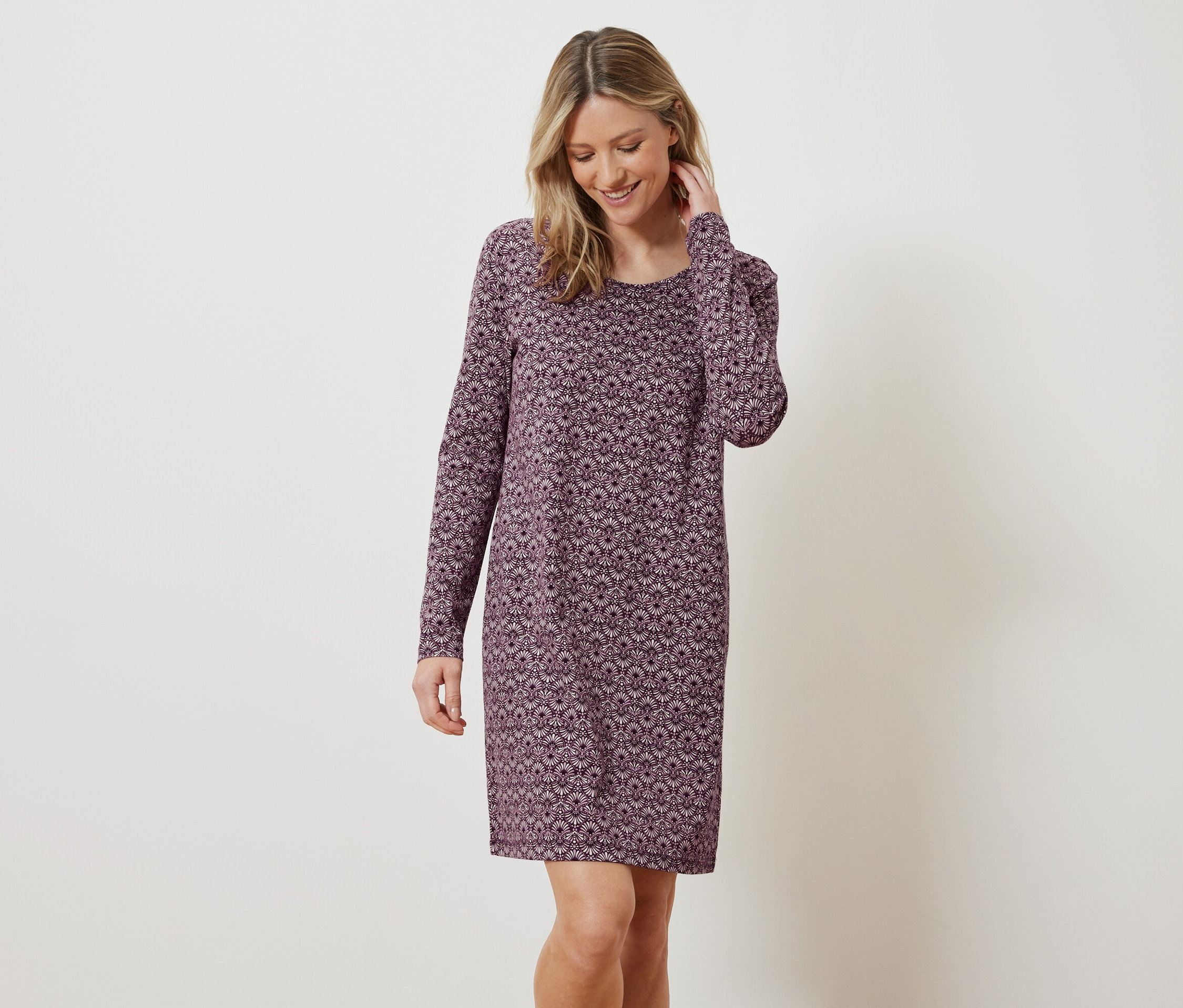 Eine blonde Frau lächelt und posiert in einem langärmligen Kleid mit Blumenmuster.