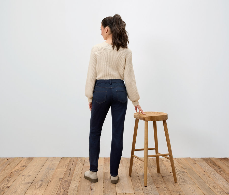 Frau mit beigem Pullover und dunkelblauen Jeans steht mit dem Rücken zur Kamera und stützt sich auf einen Holzhocker.