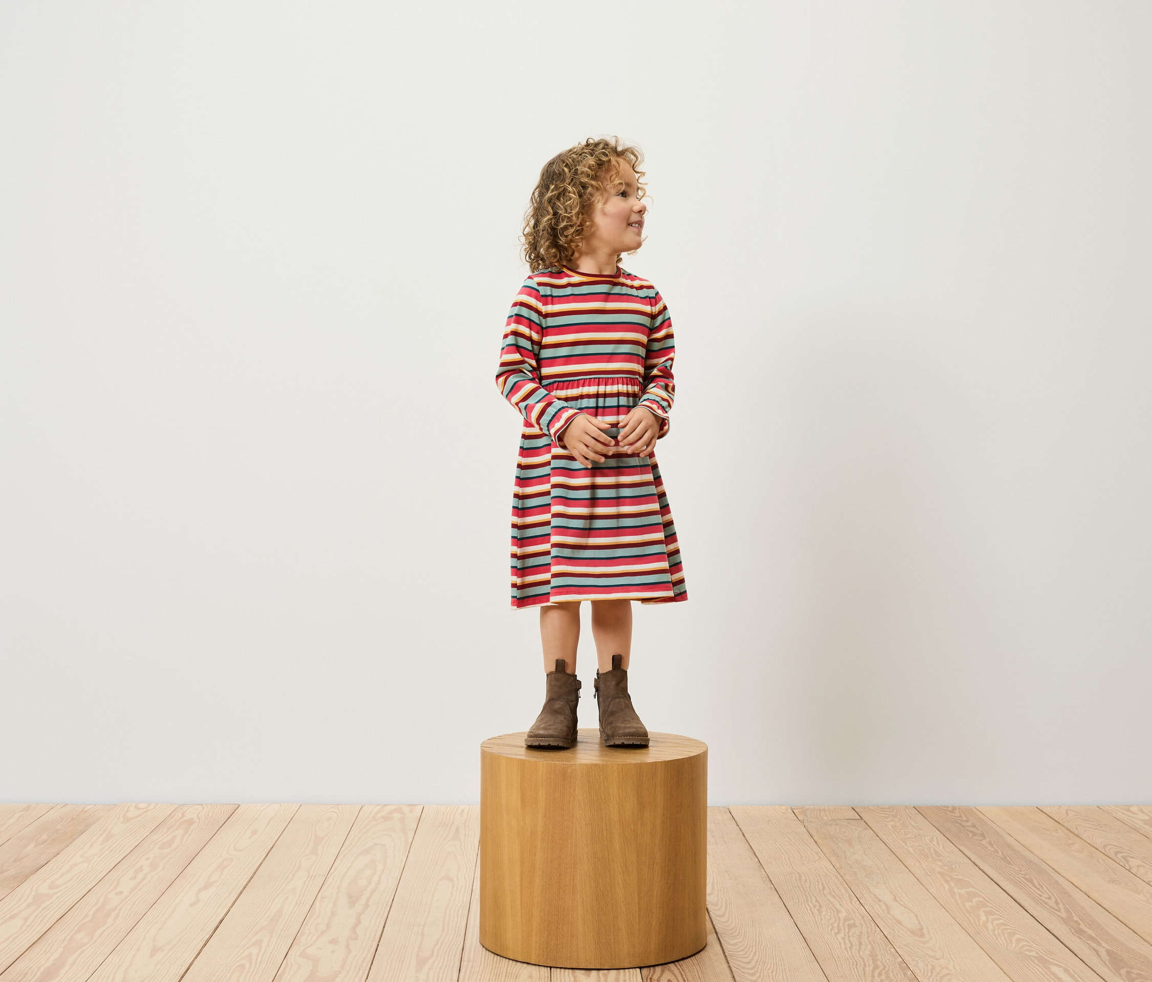 Mädchen mit lockigem Haar, bekleidet mit einem gestreiften Kleid und braunen Stiefeln, steht auf einem Holzhocker.