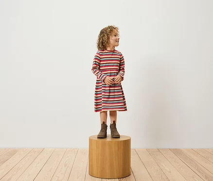 Mädchen mit lockigem Haar, bekleidet mit einem gestreiften Kleid und braunen Stiefeln, steht auf einem Holzhocker.