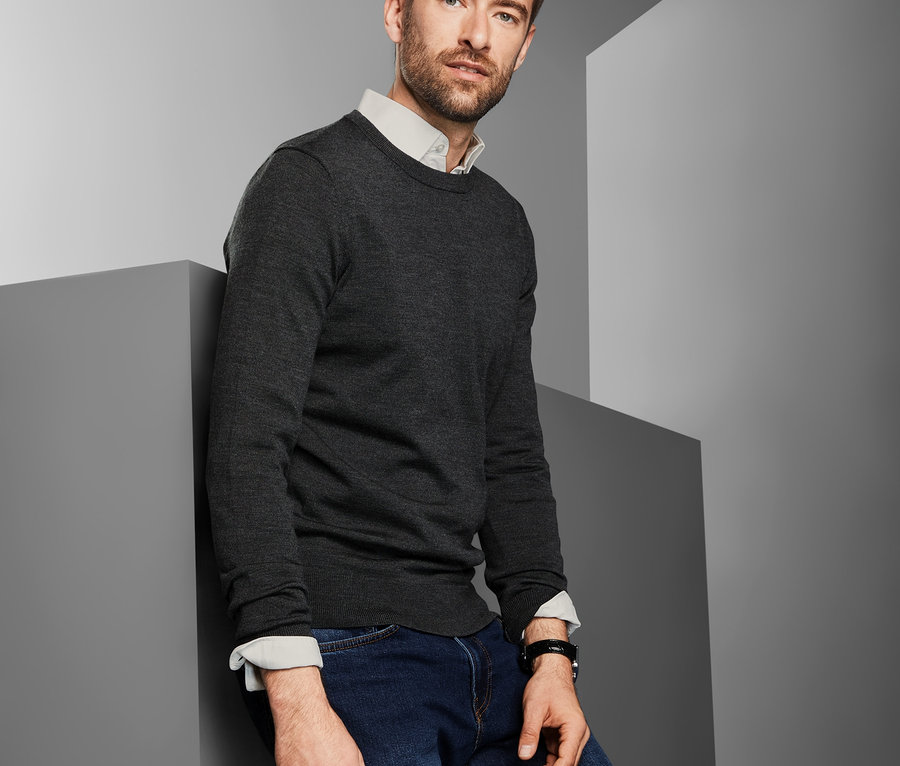 Mann lehnt an grauer Wand, trägt anthrazitfarbenen Merino-Pullover.