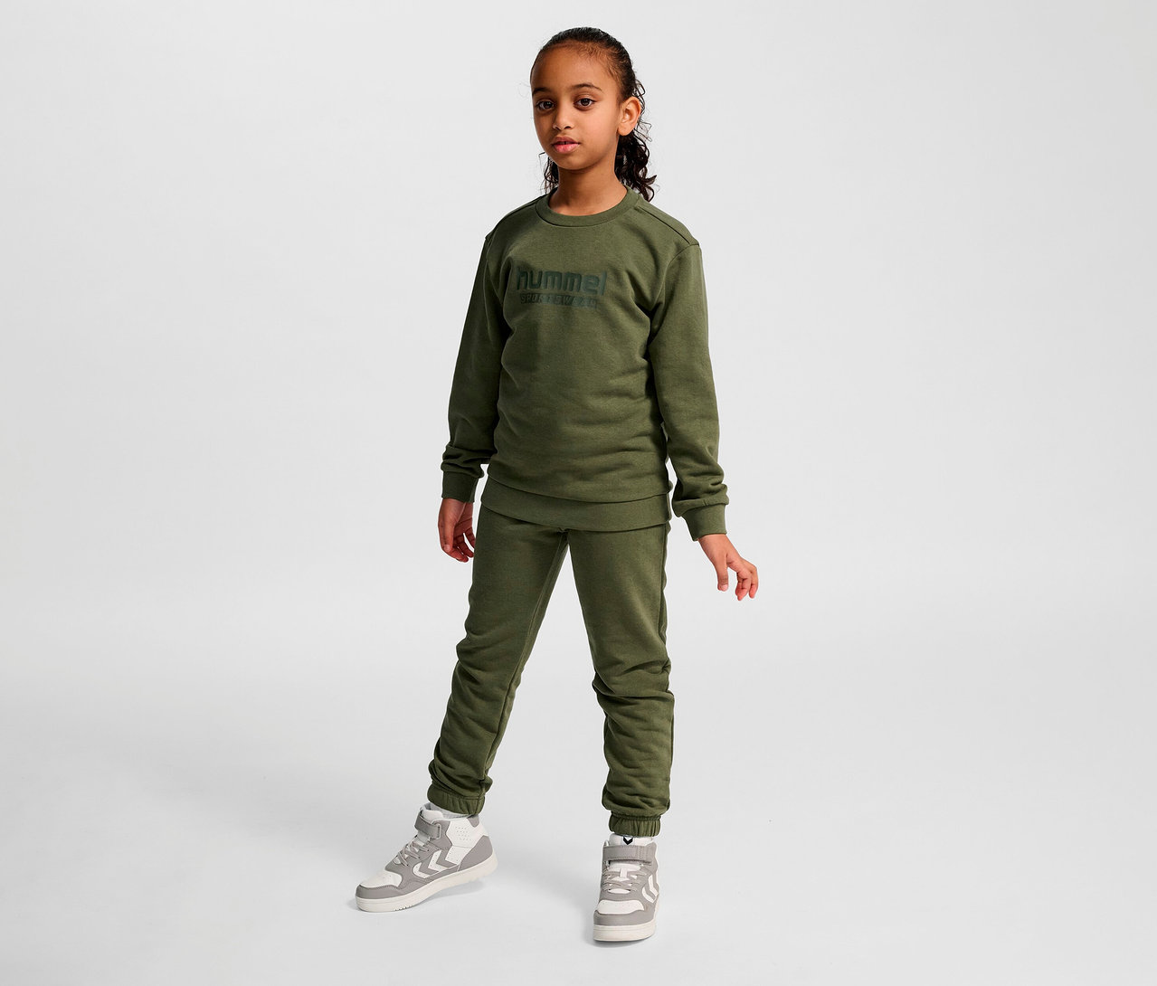 Mädchen posiert in einem grünen HUMMEL HMLJR Base-Sweatshirt und Trainingshose.