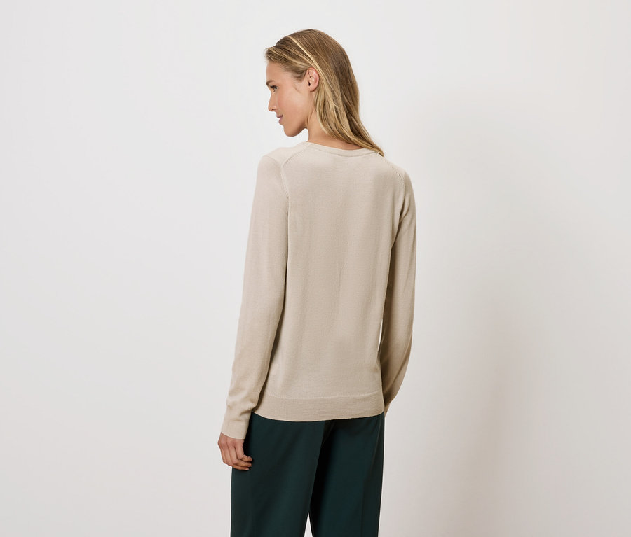 Frau blickt über die Schulter, trägt einen beigen Pullover und eine grüne Hose.