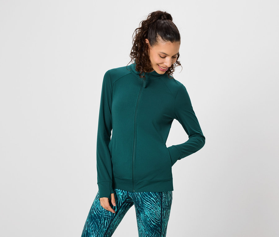 Frau mit dunklen, lockigen Haaren trägt eine grüne Trainingsjacke und gemusterte Leggings.