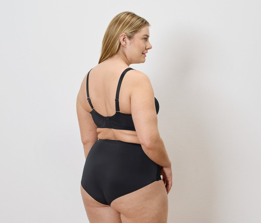 Frau mit blonden Haaren, von hinten zu sehen, trägt schwarze Unterwäsche vor einem weißen Hintergrund.