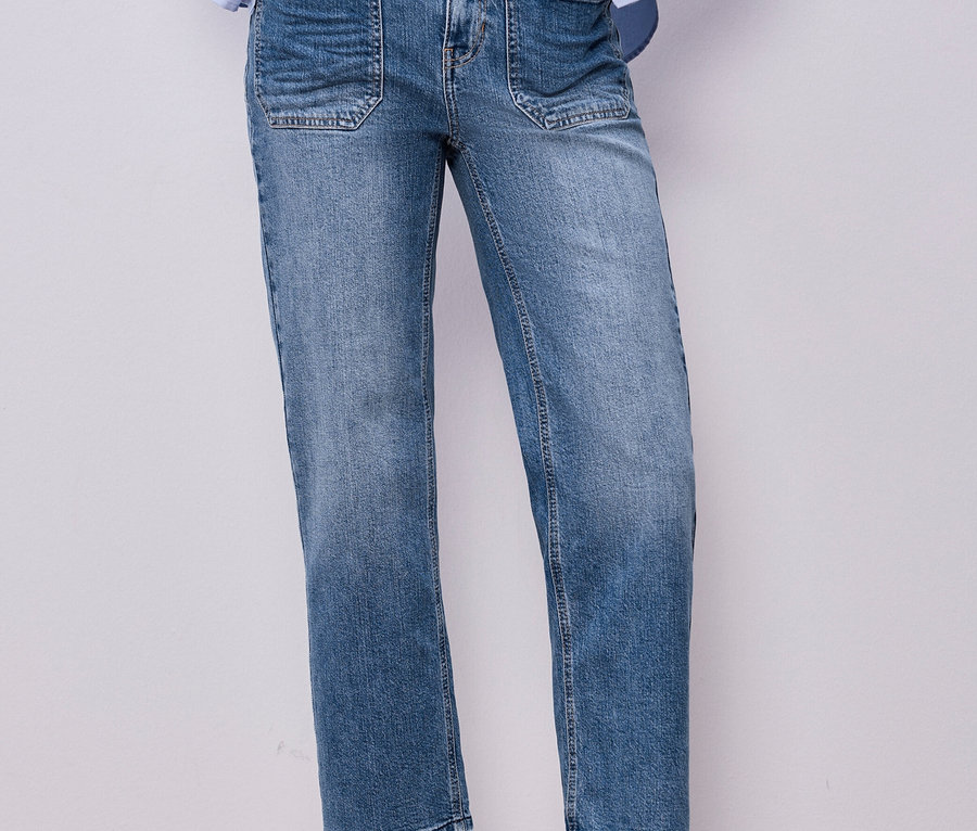Person trägt blaue Street One Jeans mit aufgesetzten Vordertaschen, ein hellblaues Hemd über einem weißen T-Shirt und braune Loafer. Hände stecken in den Hosentaschen.