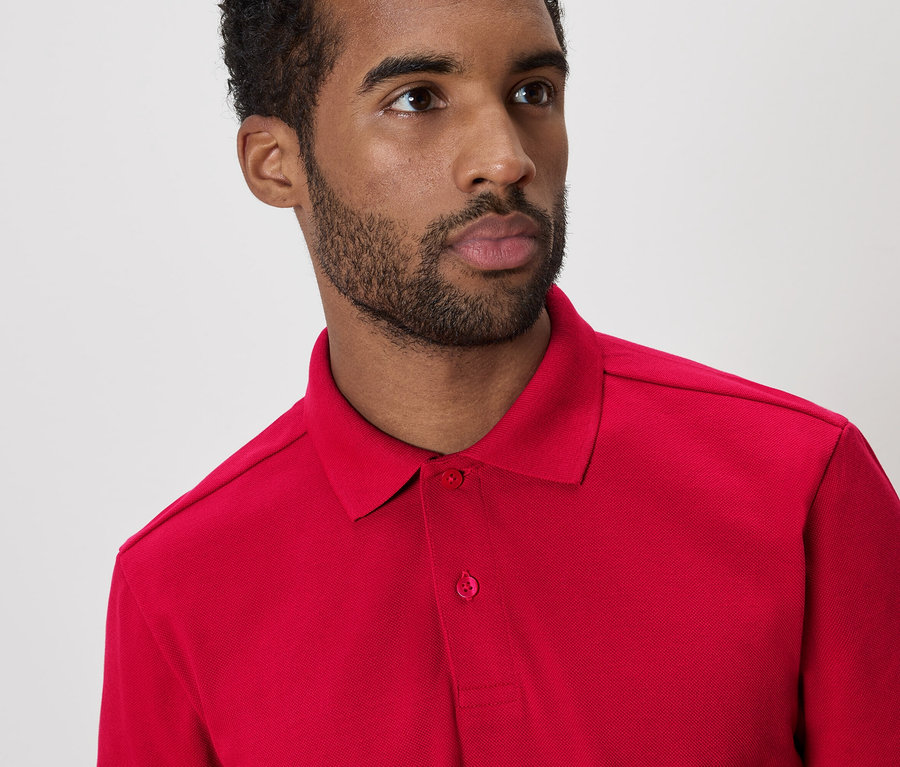 Mann mit dunklen Haaren und Bart trägt ein rotes Poloshirt vor einem weißen Hintergrund.