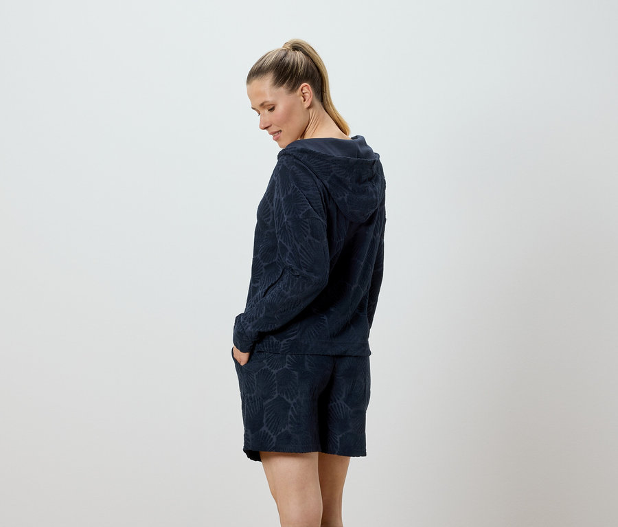 Frau mit Kapuzenpullover und Shorts mit Muschelmuster, die mit den Händen in den Taschen posiert.