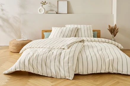 Bett mit gestreifter Bettwäsche, zwei Kissen und einer Decke. Auf dem Nachttisch stehen eine Vase und Gläser mit Wasser.