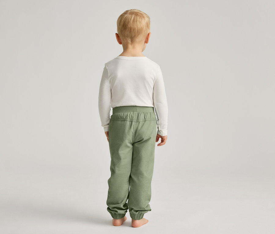 Ein Kind mit blonden Haaren steht mit dem Rücken zum Betrachter und trägt ein weißes Langarmshirt und grüne Hosen.
