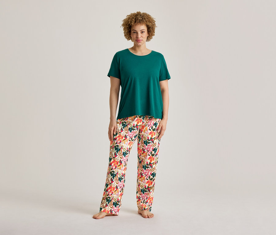 Eine Frau mit lockigem Haar steht da und trägt ein grünes T-Shirt und eine Pyjamahose mit Blumenmuster aus einem Pyjama-Set, barfuß.