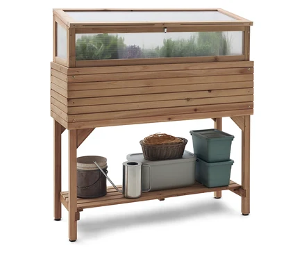 Hochbeet aus Holz mit transparentem Deckel und Ablagefläche, auf der sich eine Gießkanne, ein Korb und Behälter befinden.