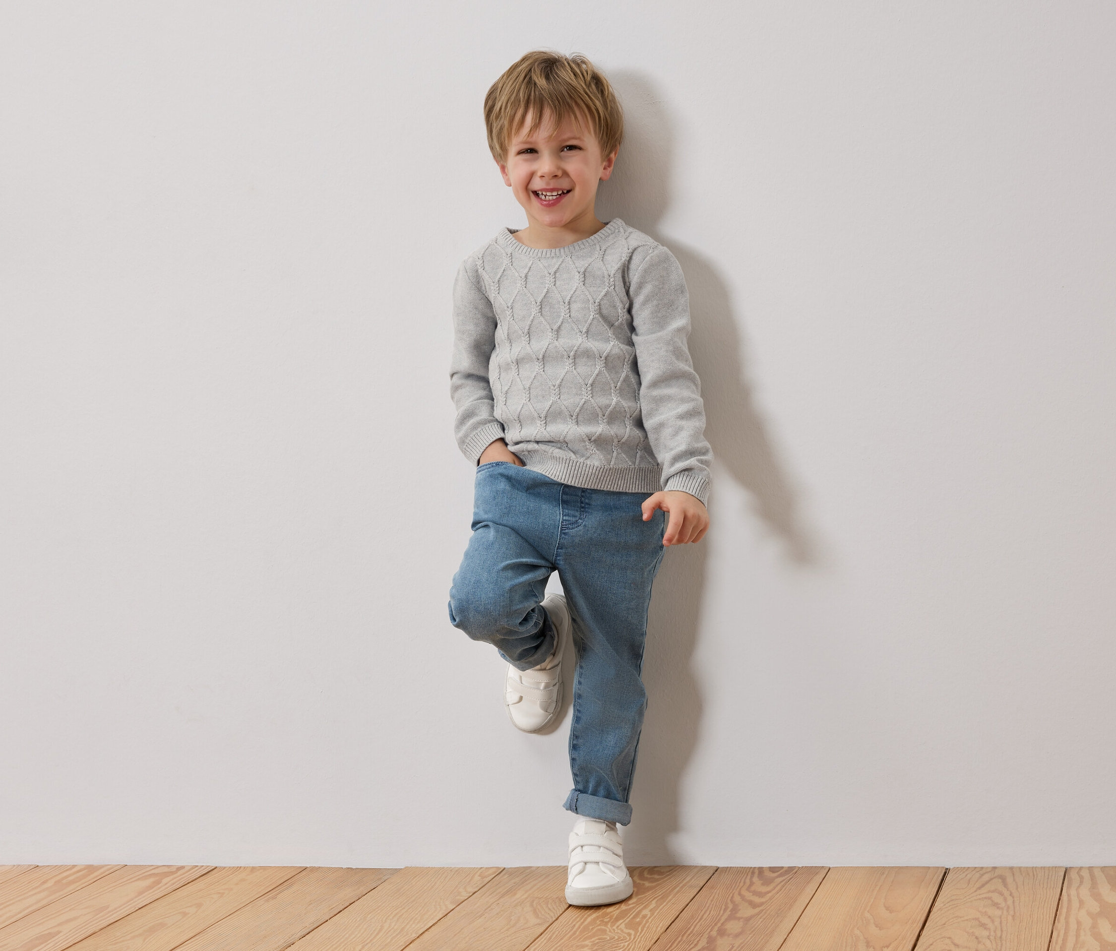 Ein lächelnder Junge mit blonden Haaren lehnt an einer weißen Wand. Er trägt einen grauen Pullover, blaue Jeans und weiße Schuhe.