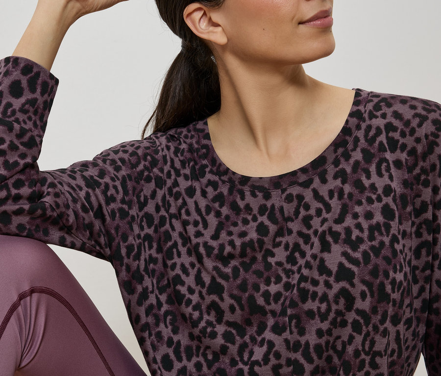 Frau sitzt und trägt ein langärmliges Oberteil mit Leopardenmuster und Leggings in Pflaumenfarbe.
