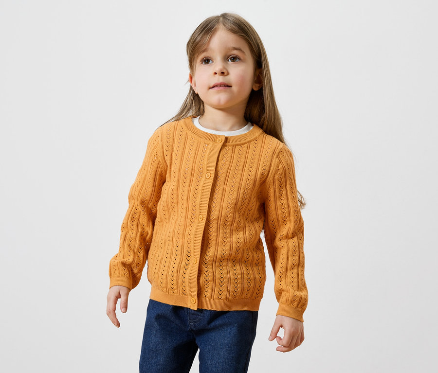 Ein Mädchen mit langen, hellbraunen Haaren trägt eine goldene Strickjacke und Jeans.
