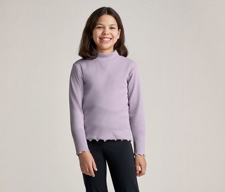 Lächelndes Mädchen mit braunen Haaren trägt ein lila Langarmshirt mit Rollkragen und schwarze Leggings vor einem hellen Hintergrund.