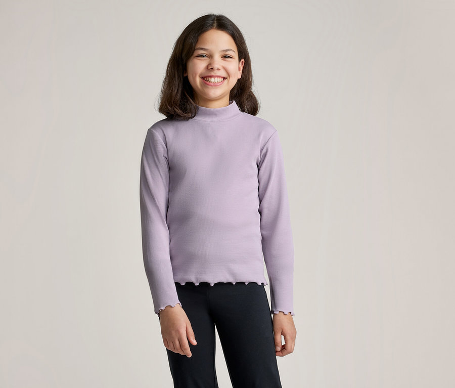 Lächelndes Mädchen mit braunen Haaren trägt ein lila Langarmshirt mit Rollkragen und schwarze Leggings vor einem hellen Hintergrund.
