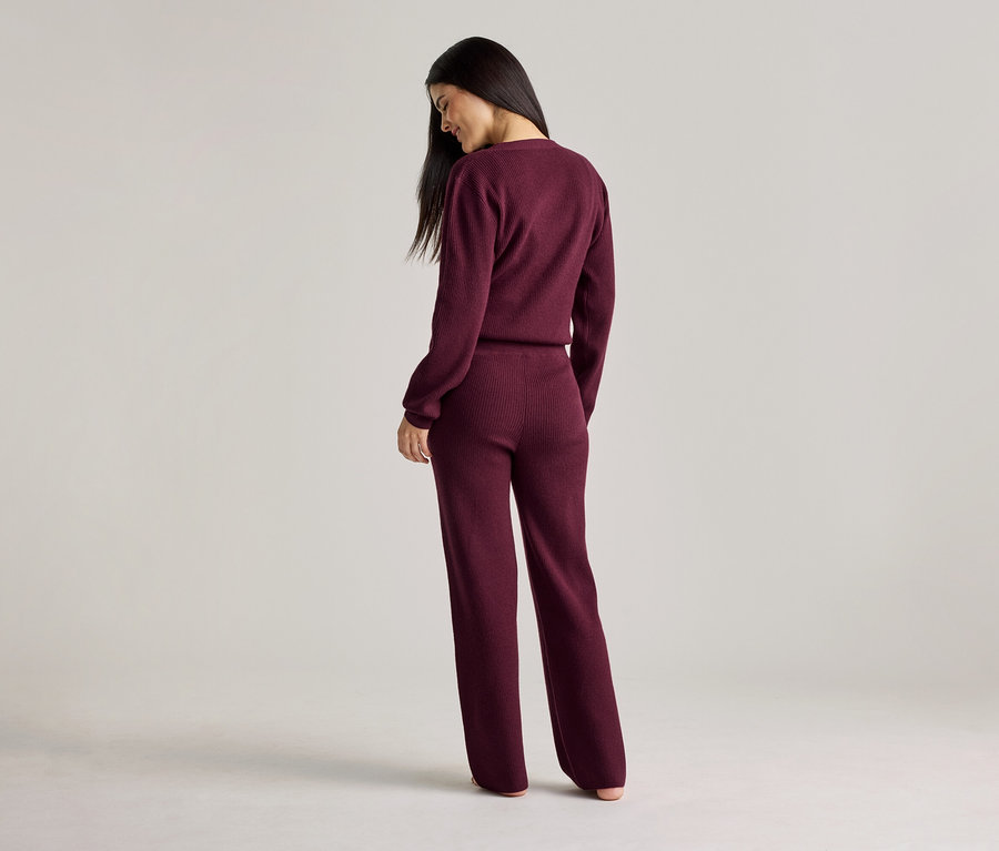 Eine Frau mit langen, dunklen Haaren steht mit dem Rücken zur Kamera und trägt ein weinrotes, geripptes Strickoberteil und eine Hose.