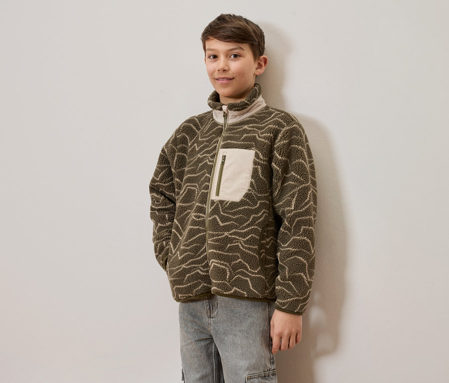 Ein junger Junge trägt eine olivgrüne Fleecejacke mit hellen Wellenmustern und hellen Jeans, steht vor einer hellen Wand. Die Jacke hat einen durchgehenden Reißverschluss und eine helle Brusttasche mit Reißverschluss.
