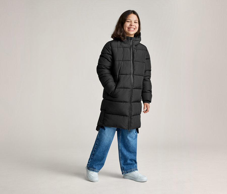 Ein lächelnder junger Mensch trägt einen langen schwarzen Steppmantel, blaue Jeans und hellgraue Sneaker und steht mit den Händen in den Taschen.
