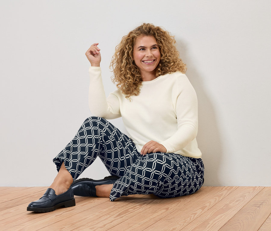 Frau mit lockigem Haar sitzt auf einem Holzboden, trägt einen cremefarbenen Pullover und eine blaue Hose mit geometrischem Muster.