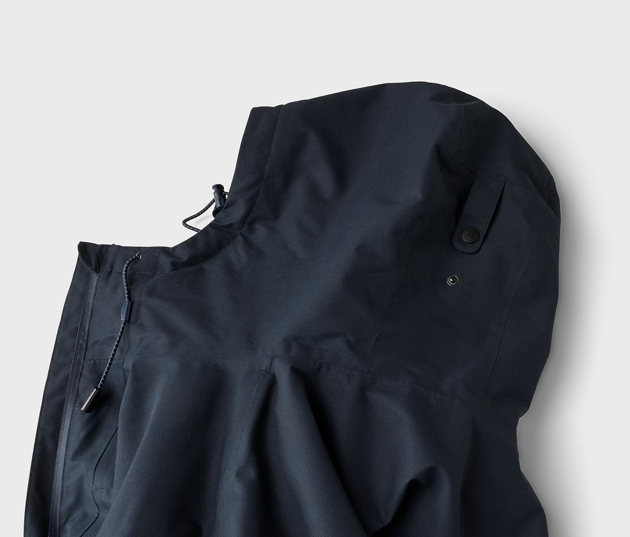 Nahaufnahme einer dunkelblauen Kapuzenjacke mit Kordelzug und Stopper am Kragen sowie zwei schwarzen Druckknöpfen an der Seite der Kapuze.
