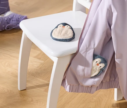 Eine hellviolette Jacke hängt auf einem weißen Stuhl, mit einem flauschigen Pinguin-Spielzeug auf der Sitzfläche und einem weiteren in der Jackentasche. Zwei lila Kinderstiefel stehen auf dem Holzboden.