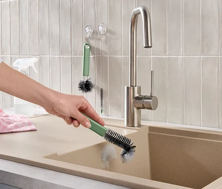 Hand reinigt beiges Spülbecken mit grüner Bürste. Ein Edelstahl-Wasserhahn, helle Wandfliesen, ein rosa Handtuch und weitere Reinigungsbürsten sind sichtbar.