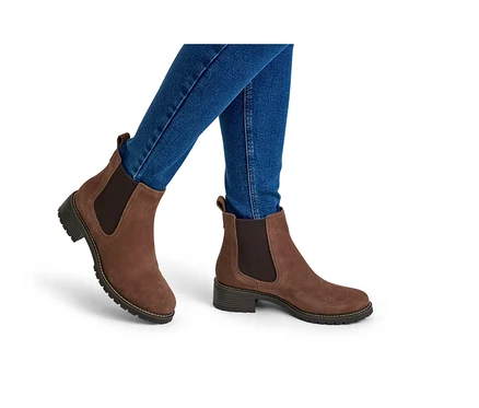 Eine Person trägt blaue Jeans und braune Wildleder-Chelsea-Boots mit elastischen Seiteneinsätzen und einer klobigen Sohle auf weißem Hintergrund.