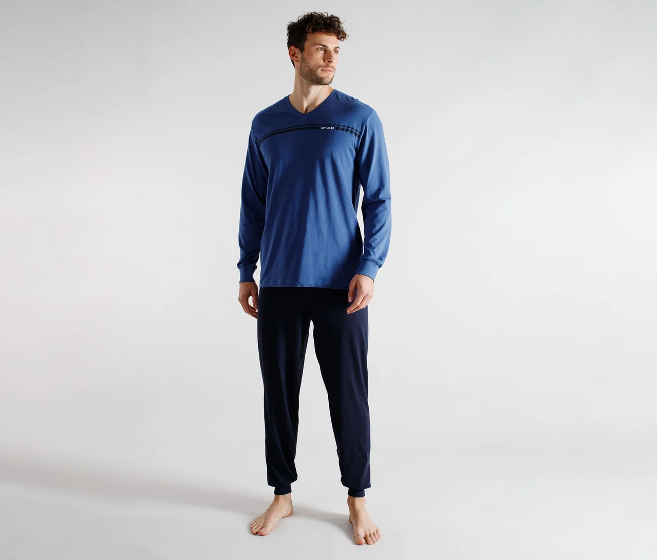 Ein Mann in einem blauen Langarm-V-Ausschnitt-Shirt mit horizontalem Streifendetail und dunkelblauer Jogginghose, barfuß stehend. Er blickt nach rechts.