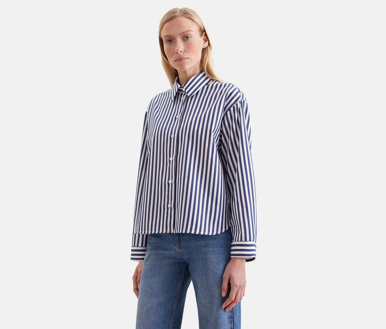 Blonde Frau in einem blau-weiß gestreiften Langarmhemd und blauen Jeans, blickt nach vorne.