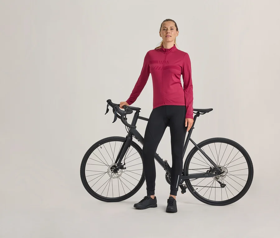 Frau steht neben einem schwarzen Rennrad, trägt ein magentafarbenes langärmeliges Radtrikot, schwarze Radhose und schwarze Schuhe.