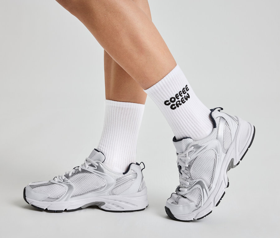 Beine einer Person in weißen Socken mit der Aufschrift "Coffee Crew" und weißen und silbernen Turnschuhen.