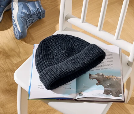 Eine dunkelblaue Strickmütze liegt auf einem aufgeschlagenen Kinderbuch, das eine Bärenillustration und Text zeigt. Das Buch liegt auf einem weißen Holzstuhl, mit zwei blauen Wanderschuhen auf dem Holzboden im Hintergrund.
