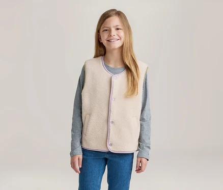 Junges Mädchen mit langen blonden Haaren lächelt und trägt eine cremefarbene Fleeceweste mit lila Besatz über einem grauen Langarmshirt und blauen Jeans.