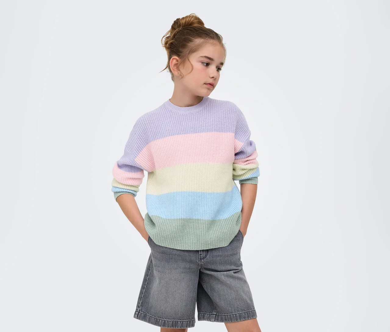 Mädchen mit Dutt trägt einen gestreiften ONLY GIRLS Sandy Strickpullover in Lavendel, Rosa, Gelb, Hellblau und Hellgrün sowie graue Jeans-Shorts, Hände in den Taschen, blickt zur Seite vor weißem Hintergrund.
