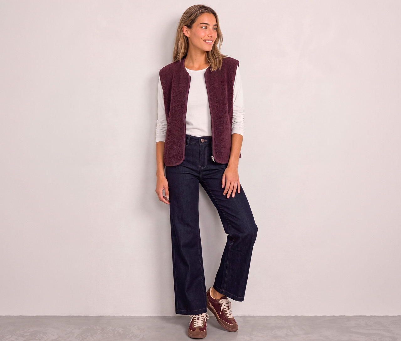 Eine lächelnde Frau trägt eine rote Street One Weste über einem weißen Langarmshirt, dazu weite Jeans und braune Sneaker.