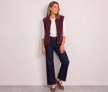 Eine lächelnde Frau trägt eine rote Street One Weste über einem weißen Langarmshirt, dazu weite Jeans und braune Sneaker.