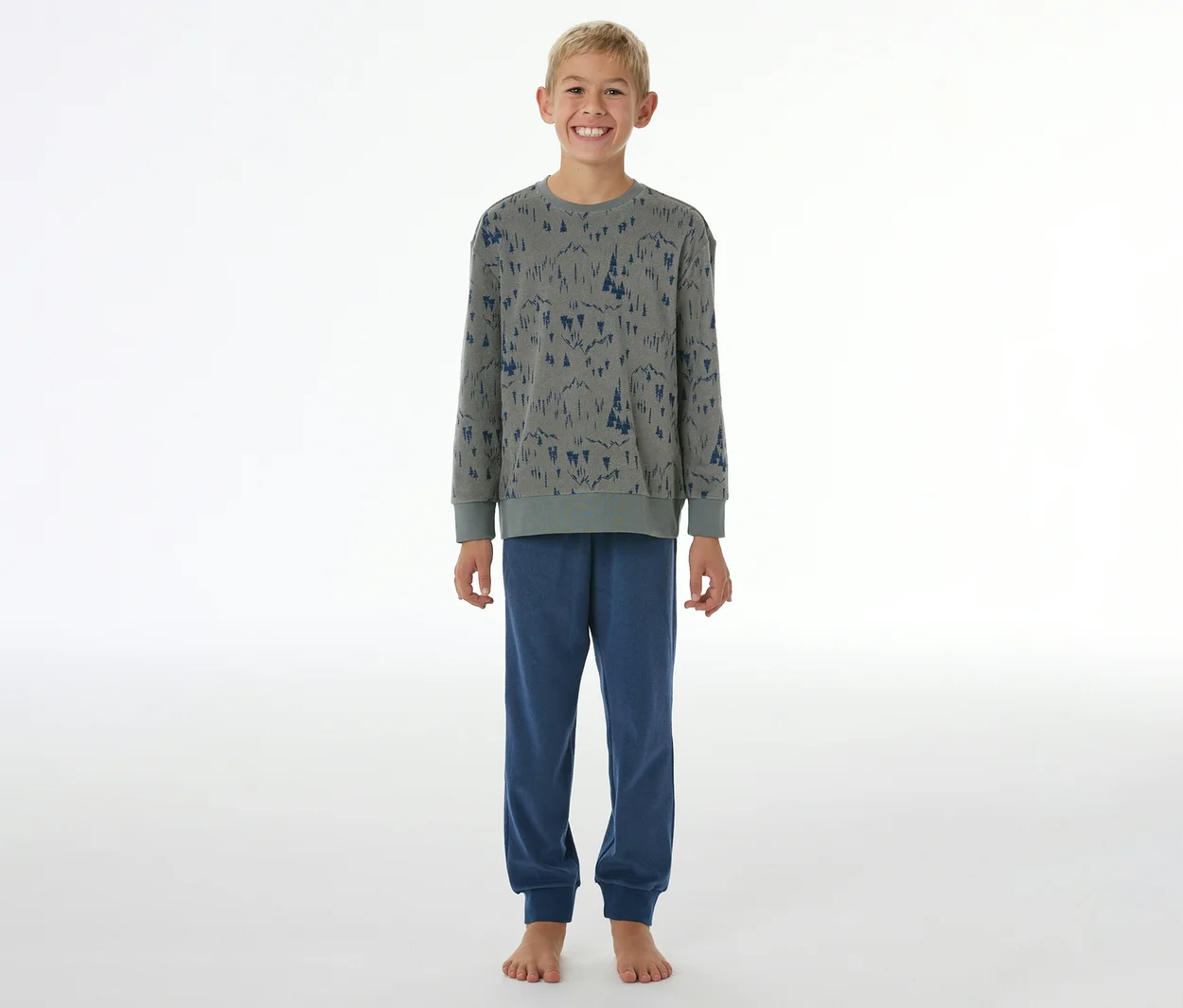 Lächelnder Junge in einem grauen SCHIESSER Kinder-Pyjama mit blauem Waldmuster und blauen Pyjamahosen mit Bündchen, barfuß stehend.