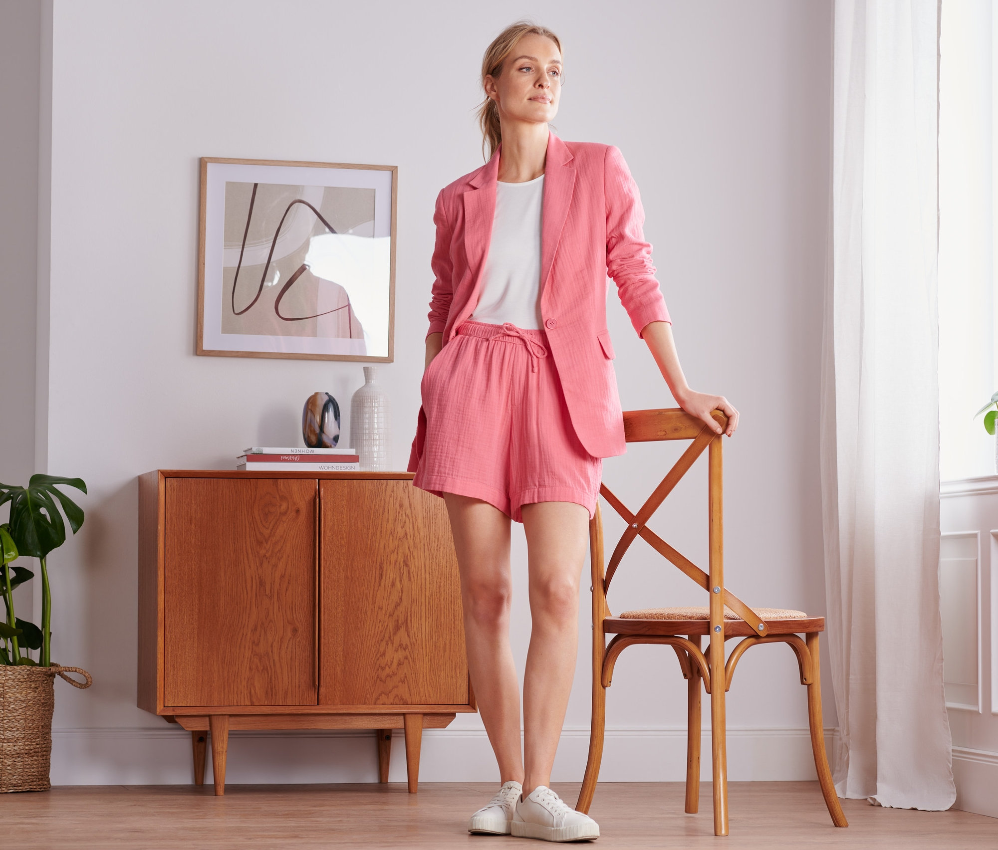 Frau posiert in korallefarbenem Musselin-Blazer und Shorts, lehnt an einem Stuhl neben einem Fenster.
