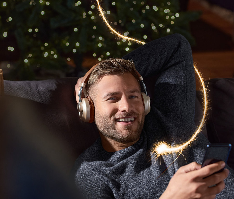 Lächelnder Mann liegt mit On-Ear-Bluetooth®-Kopfhörer auf Sofa, hält Handy in der Hand. Im Hintergrund ein Weihnachtsbaum.