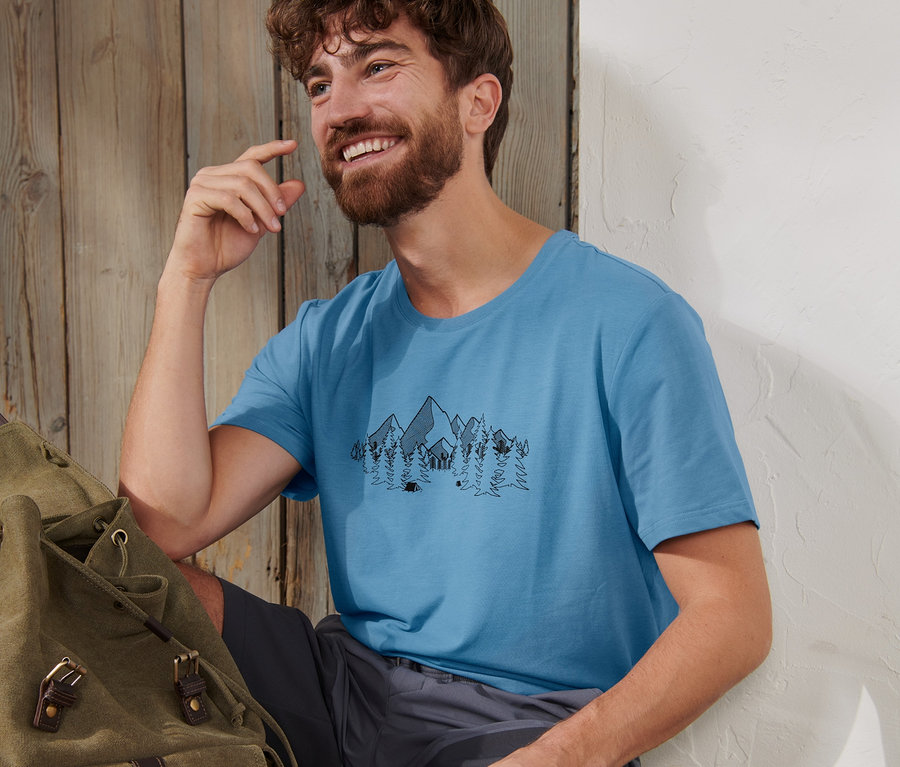 Mann sitzt neben Rucksack und Jause auf Holzboden und trägt blaues Funktionsshirt.