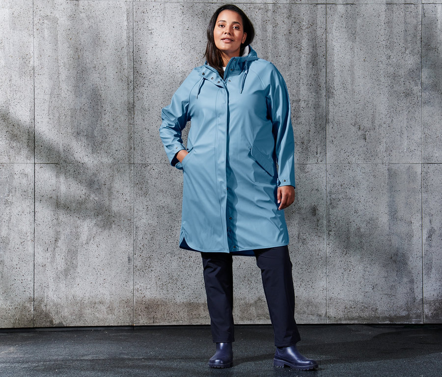 Frau steht und trägt einen hellblauen Regenparka »Friesennerz« und eine dunkelblaue Softshell-Joggpants.