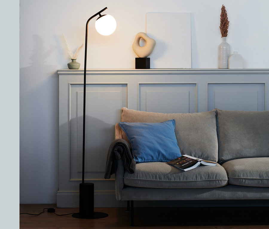 Eine schwarze Stehleuchte mit Glaskugel steht neben einem grauen Sofa.