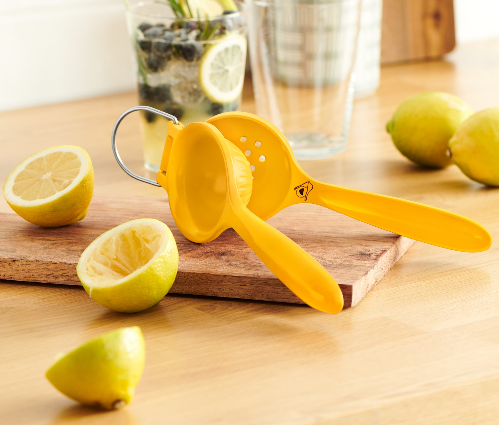 Gelbe Zitruspresse auf einem Holzbrett neben Zitronen und einem Glas Zitronenwasser.