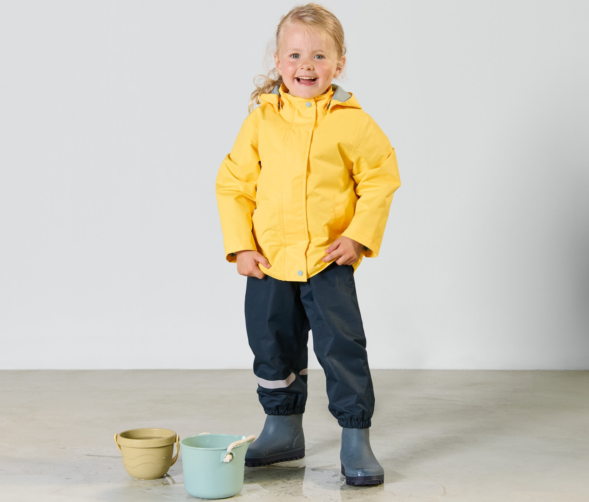 Ein lächelndes kleines Mädchen steht in einer gelben 3-in-1-Kinder-Outdoorjacke und dunklen Hosen mit Gummistiefeln neben zwei Eimern.
