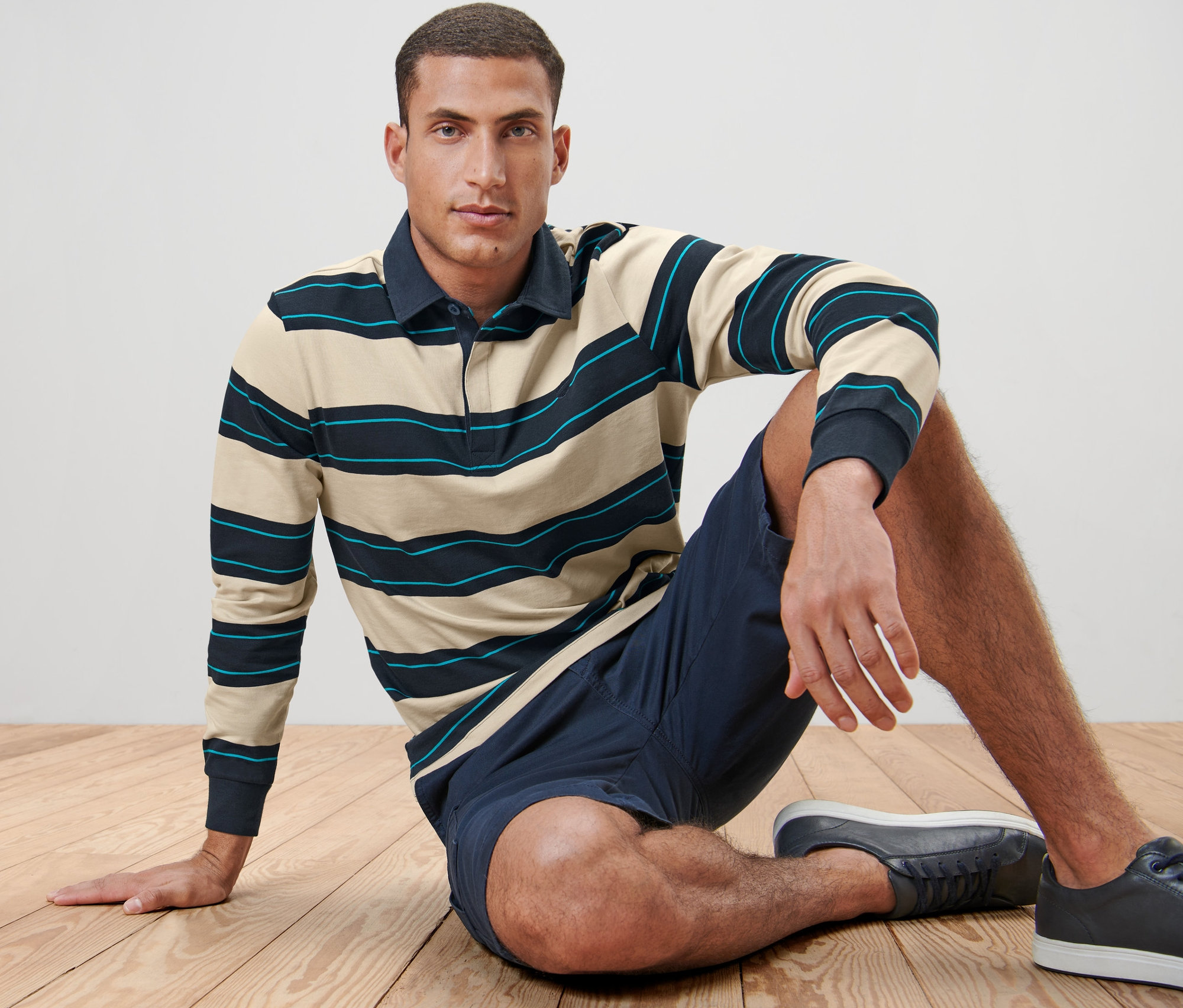 Mann sitzt auf Holzboden und trägt ein Langarm-Poloshirt, Shorts und Turnschuhe.
