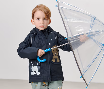 Junge hält durchsichtigen Regenschirm und trägt Kinder-Thermo-Regenjacke, Hund.